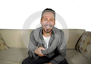 Young attractive and happy man laughing at home watching television show or comedy series holding remote sitting at living room