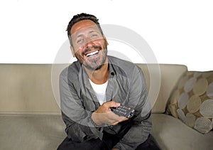 Young attractive and happy man laughing at home watching television show or comedy series holding remote sitting at living room
