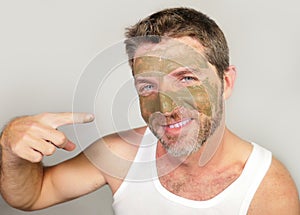 Young attractive and happy man laughing looking at himself in the mirror funny with green cream on his face applying facial mask