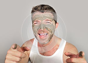 Young attractive and happy man laughing looking at himself in the mirror funny with green cream on his face applying facial mask