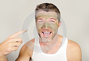 Young attractive and happy man laughing looking at himself in the mirror funny with green cream on his face applying facial mask