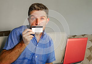 Young attractive and happy man relaxed at home sofa couch using credit card and laptop computer for internet shopping and banking