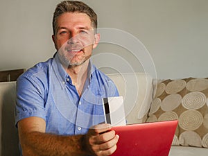 Young attractive and happy man relaxed at home sofa couch using credit card and laptop computer for internet shopping and banking