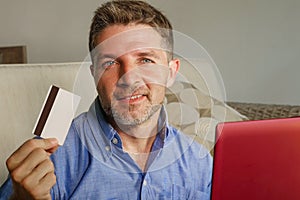 Young attractive and happy man relaxed at home sofa couch using credit card and laptop computer for internet shopping and banking