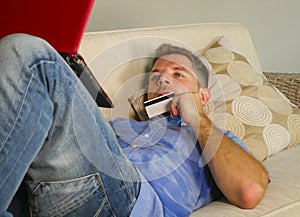 Young attractive and happy man relaxed at home sofa couch using credit card and laptop computer for internet shopping and banking