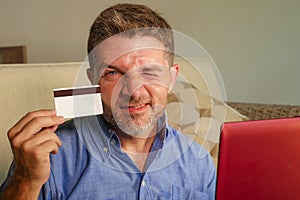 Young attractive and happy man relaxed at home sofa couch using credit card and laptop computer for internet shopping and banking