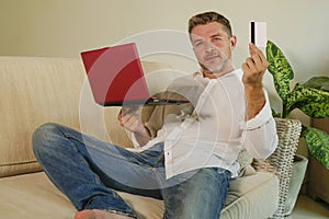 Young attractive and happy man relaxed at home sofa couch using credit card and laptop computer for internet shopping and banking