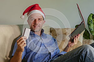 Young attractive and happy man in Santa Klaus Christmas hat using laptop computer for buying xmas presents and gifts online with c