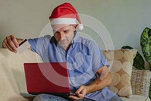 Young attractive and happy man in Santa Klaus Christmas hat using laptop computer for buying xmas presents and gifts online with c