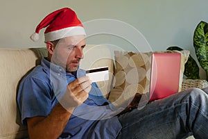 Young attractive and happy man in Santa Klaus Christmas hat using laptop computer for buying xmas presents and gifts online with c