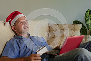 Young attractive and happy man in Santa Klaus Christmas hat using laptop computer for buying xmas presents and gifts online with c