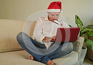 Young attractive and happy man in Santa Klaus Christmas hat using laptop computer for buying xmas presents and gifts online with c
