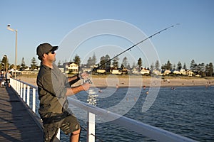 Young attractive and happy man in shirt and hat fishing at beach sea dock using fish road enjoying weekend hobby in holidays