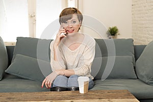 Young attractive and happy red hair woman sitting at home sofa couch drinking coffee talking on mobile phone relaxed in communicat