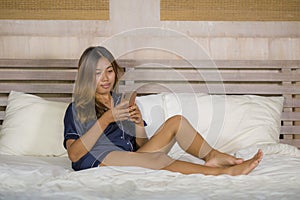 Young attractive happy and relaxed Asian teenager woman in pajamas at home bed using dating app or texting her boyfriend on mobile