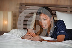 Young attractive happy and relaxed Asian teenager woman in pajamas at home bed using dating app or texting her boyfriend on mobile