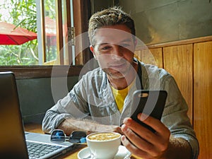 Young attractive happy and successful millennial man working from internet coffee shop with laptop computer and mobile phone relax