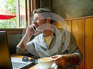 Young attractive happy and successful millennial man working from internet coffee shop with laptop computer talking on mobile phon