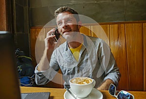 Young attractive happy and successful millennial man working from internet coffee shop with laptop computer talking on mobile phon