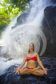 Young attractive and happy woman with fit body practicing yoga wet under tropical paradise waterfall stream sitting in lotus pose