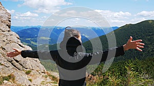 A young attractive hiker woman stands on the top of a mountain with her arms outstretched to the sides, enjoying the