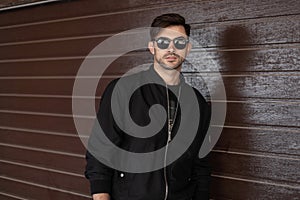 Young attractive hipster man in stylish black jacket in trendy sunglasses with fashionable hairstyle relaxes near brown vintage