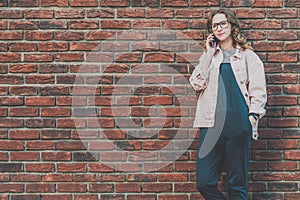 Young attractive hipster woman stands outside on a red brick wall background and talks on her cell phone.