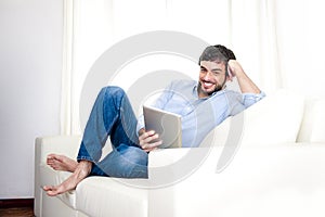 Young attractive Hispanic man at home on white couch using digital tablet or pad