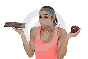 Young attractive hispanic woman in fitness top holding apple fruit and chocolate bar in her hands