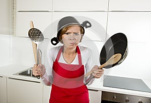 Young attractive home cook woman in red apron at kitchen holding pan and household with pot on her head in stress