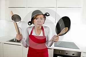 Young attractive home cook woman in red apron at kitchen holding pan and household with pot on her head in stress