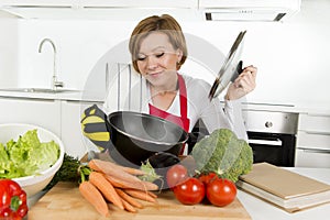 Young attractive home cook woman in red apron at kitchen holding pan and household with pot on her head in stress