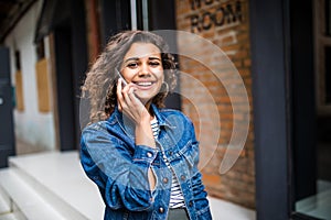 Young attractive latin curly woman stands on city street and talking on her cell phone on background red brick wall. Girl is talki