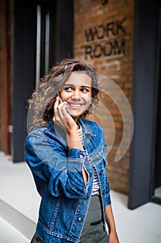 Young attractive latin curly woman stands on city street and talking on her cell phone on background red brick wall. Girl is talki