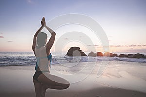 Young attractive latin woman wearing a bikini practicing yoga on the beach at sunset, healthy mental and body lifestyle concept