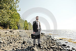 Young attractive male businessman in a brown suit with a briefcase stands on the seashore. Unfastens shirt, loosens tie