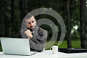 Young attractive male businessman in brown suit and tie sits at desk and works on computer outdoors. Green trees, nature