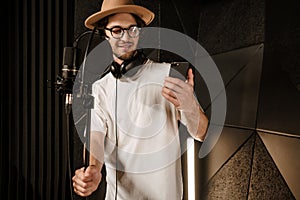 Young attractive male musician reading song text on smartphone preparing singing in sound recording studio