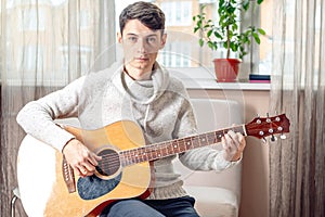 Young attractive male musician sitting on a chair playing acoustic guitar in room. Concept of music as a hobby