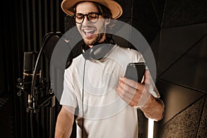 Young attractive male singer with smartphone emotionally singing in recording studio