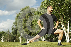 Young attractive man in black sportwear doing lunge outdoor in the park.