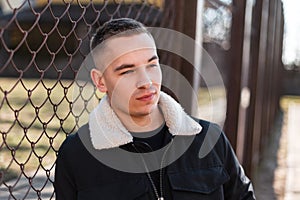 Young attractive man in a black stylish jacket with a white collar in a trendy T-shirt is rest standing near a metal vintage fence