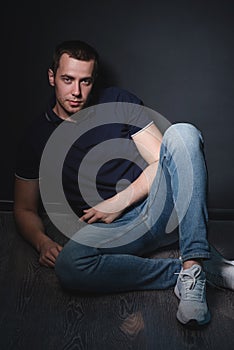 Young attractive man in blue jeans and a polo shirt sits on the floor near the wall in the studio. Youth and