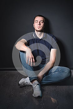 Young attractive man in blue jeans and a polo shirt sits on the floor near the wall in the studio. Youth and