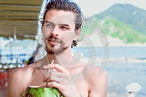 Young attractive man drinks juice of green coconut and looking away in the background of the bay and mountains.