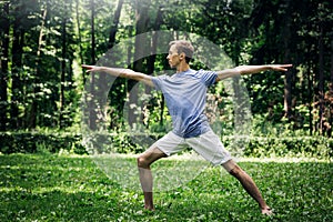 Young attractive man in a gray T-shirt and sweatpants doing yoga warrior pose in the park