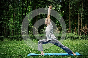 Young attractive man in a gray T-shirt and sweatpants doing yoga warrior pose in the park.