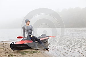 Young attractive man in grey cap and shirt, posing on bank of river, looking dreamily in distance, having rest after rowing boat,