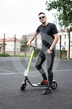 Young attractive man hipster in a black T-shirt in jeans in sneakers in sunglasses stands with a modern electric scooter