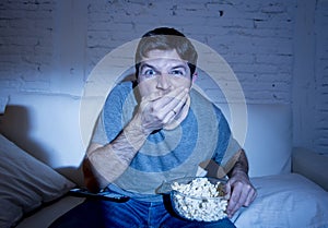 Young attractive man at home lying on couch at living room watching tv eating crudely popcorn holding bowl